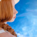 Side profile of a woman with long braided red hair wearing a sun hat by a blue pool, representing healthy summer hair care and sun protection.
