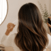 Woman with healthy, shiny hair using heat protectant before styling with a blow dryer in a modern salon-inspired bathroom, illustrating heat damage prevention.
