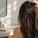 Woman massaging oily scalp roots at bathroom mirror with hair care products on marble counter